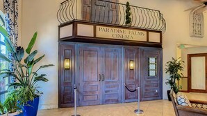 Ein Kinoeingang mit einem Schild, auf dem 'Paradise Palms Cinema' steht. Vor der Tür befinden sich Kübelpflanzen und eine Bank.