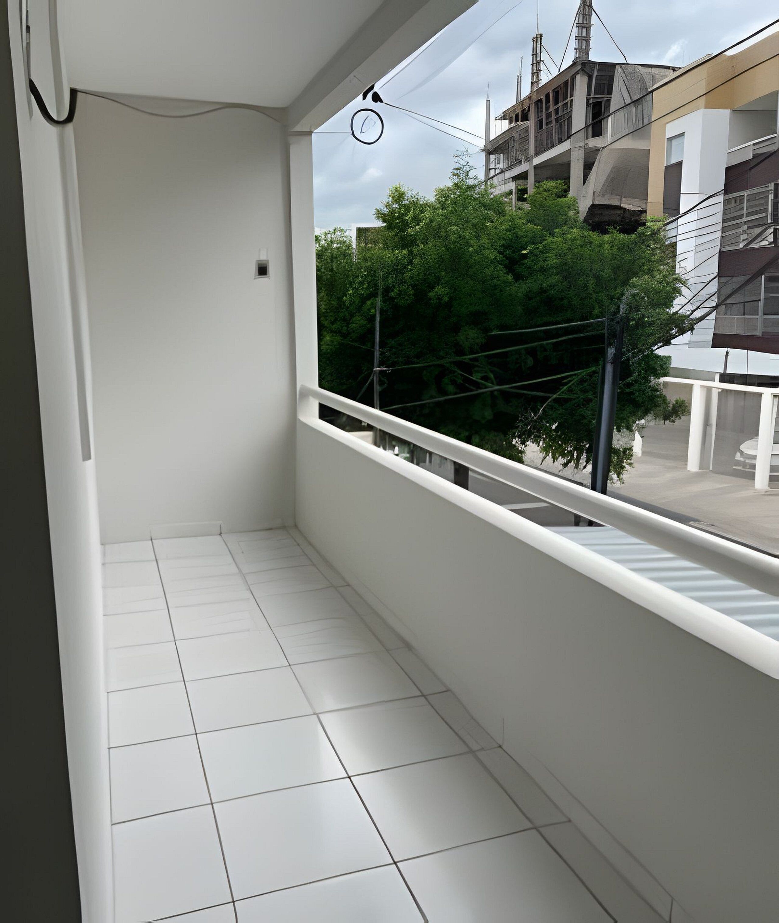 Un balcón con suelo de baldosas blancas, barandilla blanca y vistas a un edificio con balcón y vegetación.