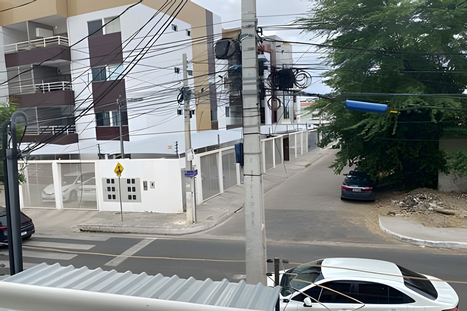 Vista de la calle con un coche blanco estacionado en la banquina, un edificio de varios pisos y un árbol al fondo.