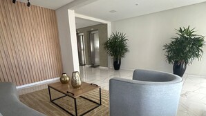 A modern lobby with a grey sofa, a wooden accent wall, a coffee table with decorative items, and two potted plants.