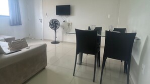 Un salon comprenant un canapé, une table à manger avec des chaises, un téléviseur fixé au mur et un ventilateur sur pied.