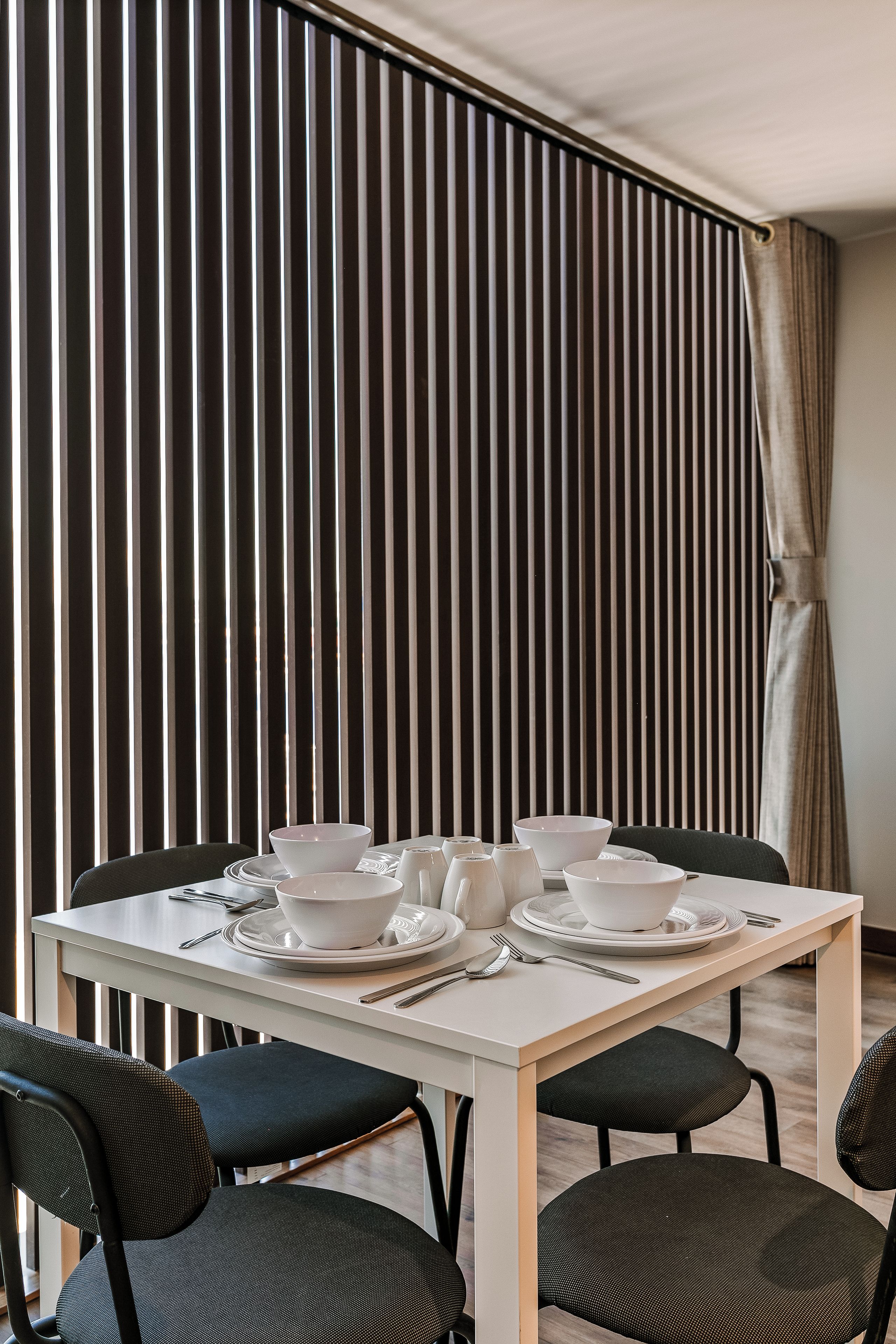 A dining area with a table set for two, chairs, and a backdrop of vertical blinds.