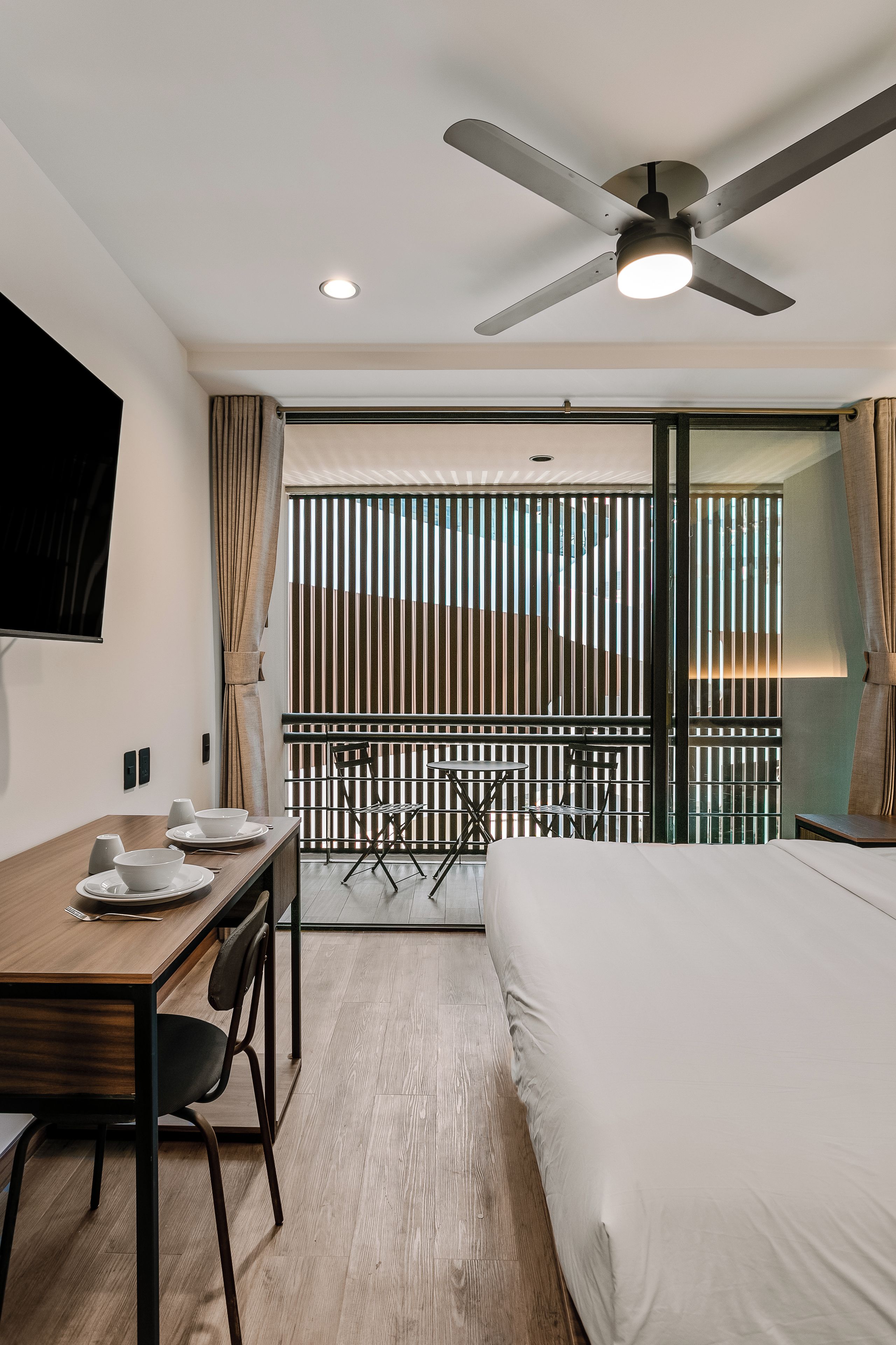 Ein Hotelzimmer mit Bett, Esstisch mit Stühlen, Deckenventilator und Blick durch ein Fenster mit Jalousien.