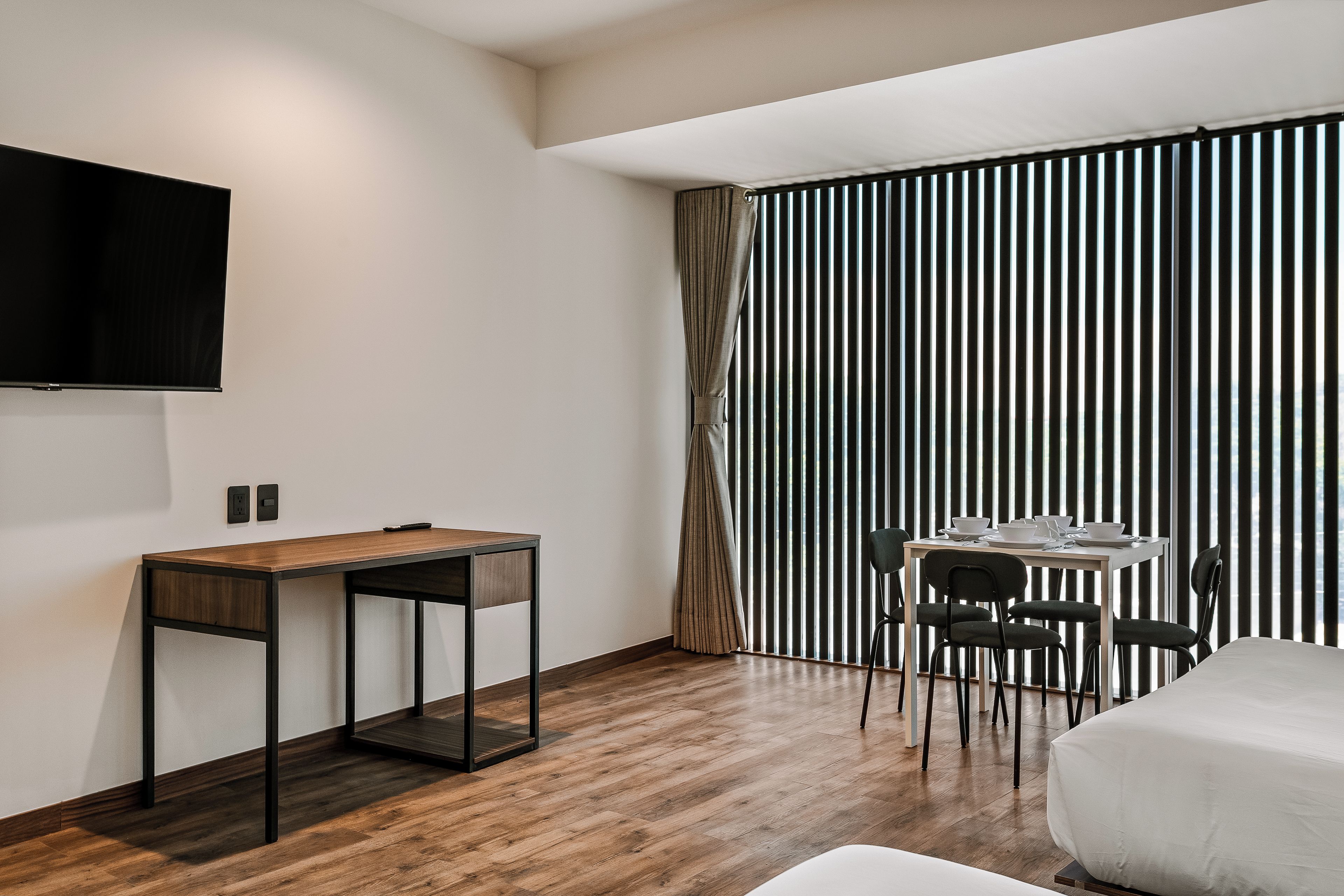 A modern hotel room with a wooden desk, a dining table with chairs, and a large window with vertical blinds.