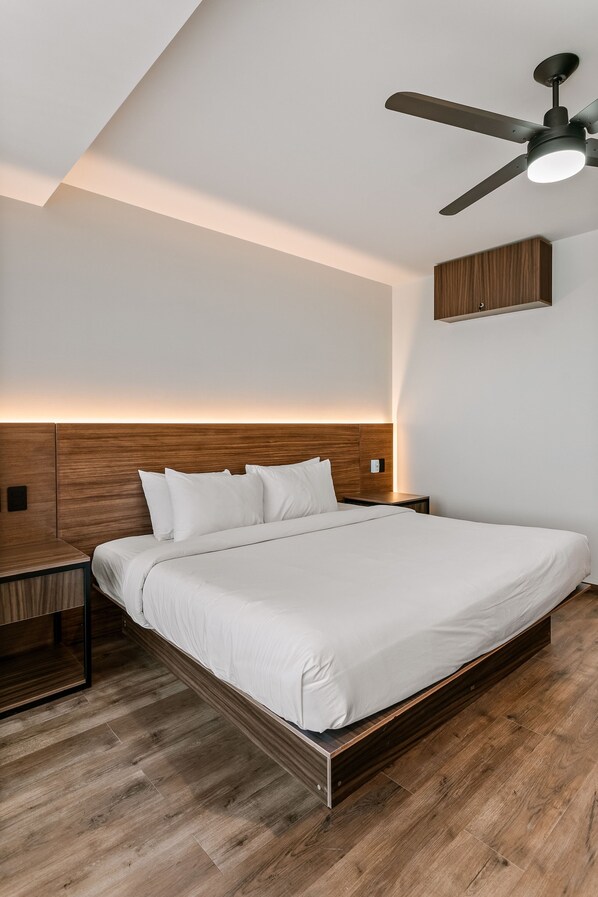 A modern hotel room with a wooden headboard, a single bed, a nightstand, a ceiling fan, and a wall-mounted cabinet. - Suites Álamos by Homirent (Querétaro)