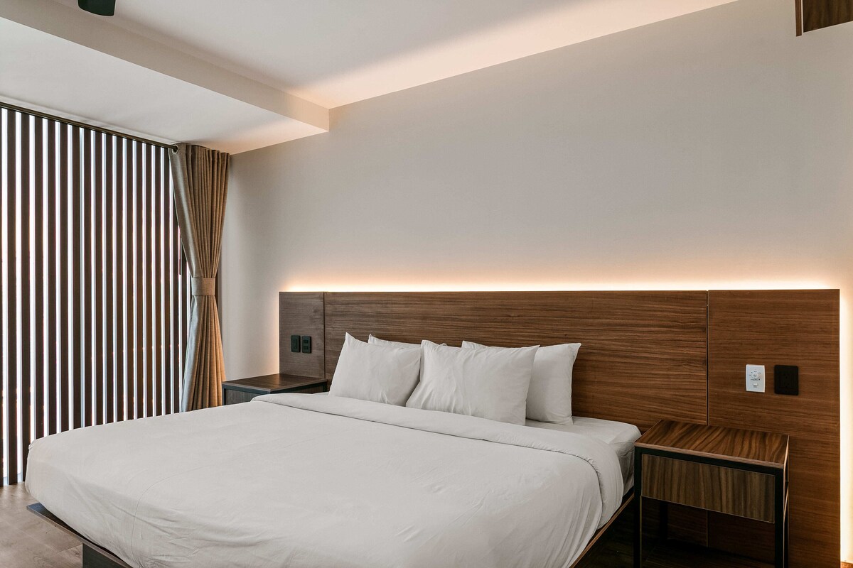 A modern hotel room with a large bed, a wooden headboard, a nightstand, and a wall-mounted control panel.
