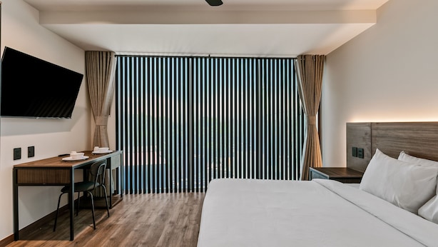 A modern hotel room with a wooden desk, a flat-screen TV, a bed with white linens, and a large window with vertical blinds. - Suites Álamos by Homirent (Querétaro)