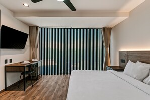 A modern hotel room with a wooden desk, a flat-screen TV, a bed with white linens, and a large window with vertical blinds. - Suites Álamos by Homirent (Querétaro)