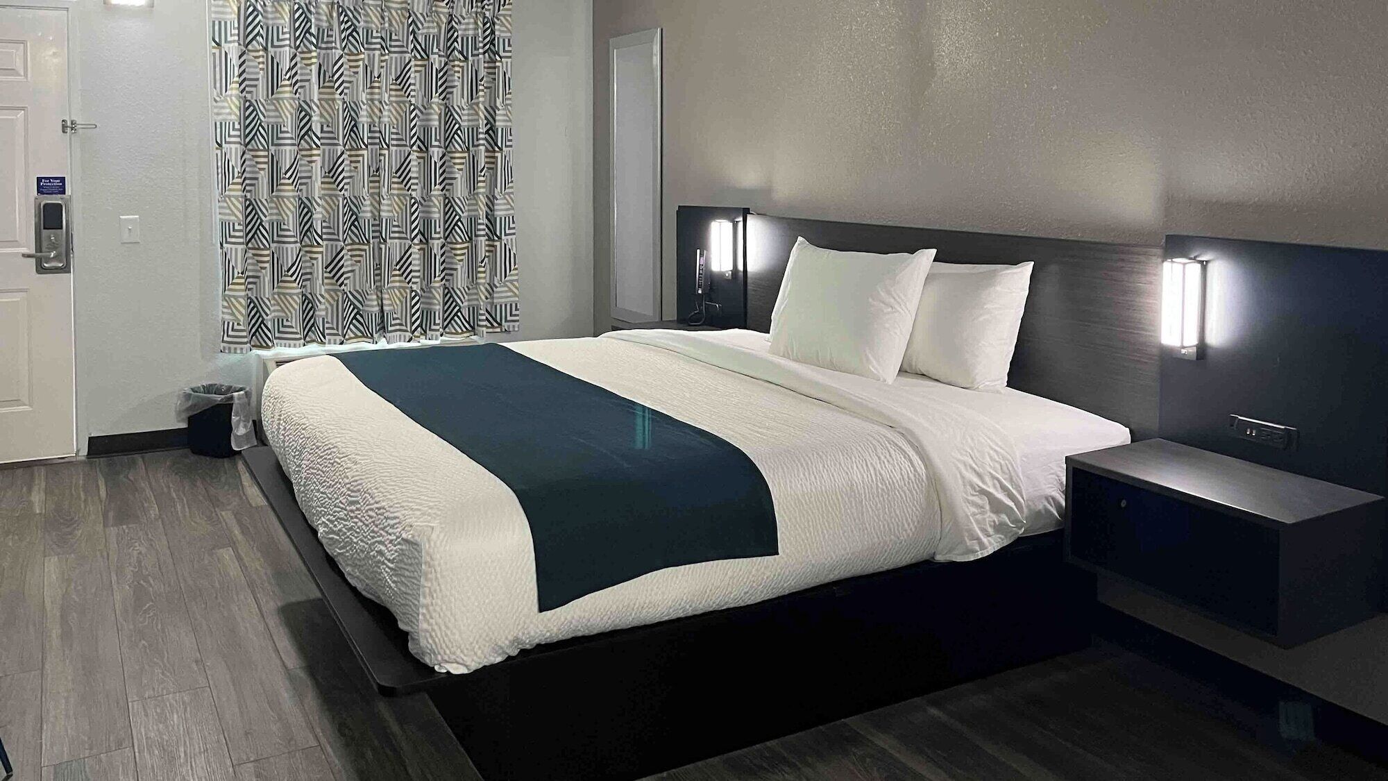 Une chambre d’hôtel moderne avec un grand lit, des tables de chevet, un rideau à motifs et un interrupteur sur le mur.