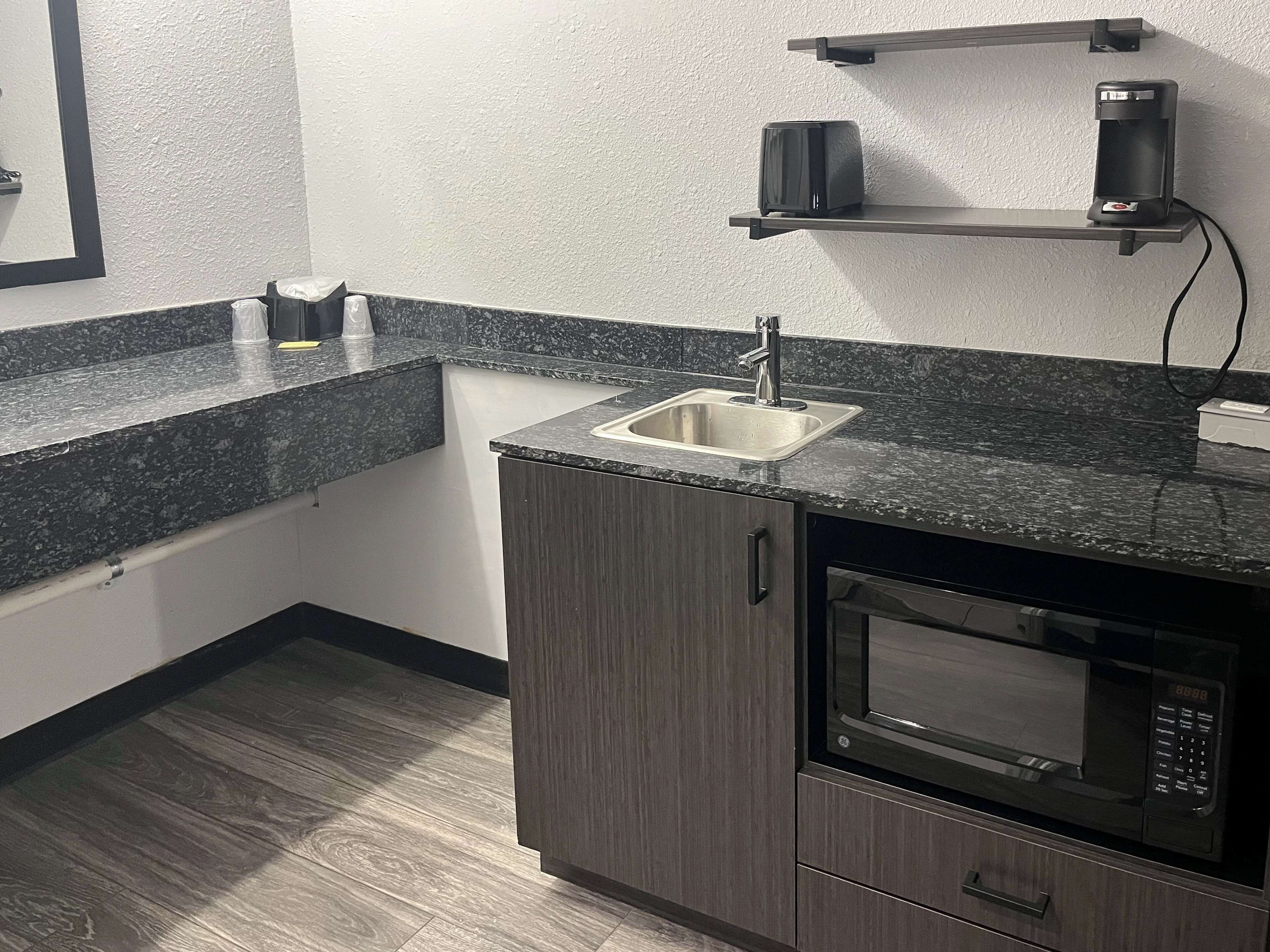 A compact kitchen with a granite countertop, a stainless steel sink, a microwave, and a coffee machine on a shelf.