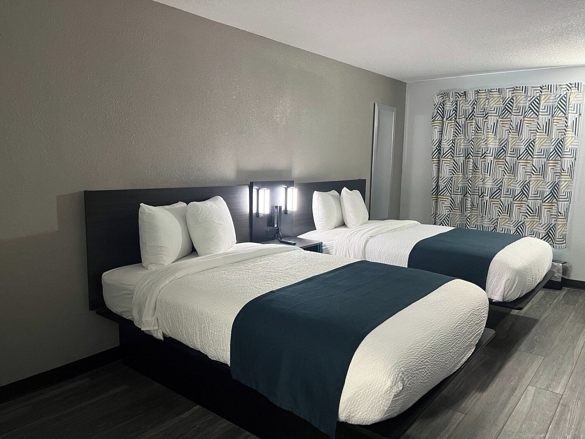A hotel room with two beds, each with white linens and dark blue bedspreads. There is a nightstand with a lamp between the beds, and a patterned curtain on the window.