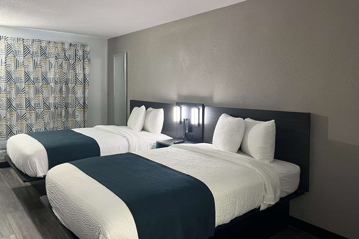 Une chambre d’hôtel avec deux lits, un rideau à motifs, une tête de lit dotée d’un panneau de commande et un plancher en bois.