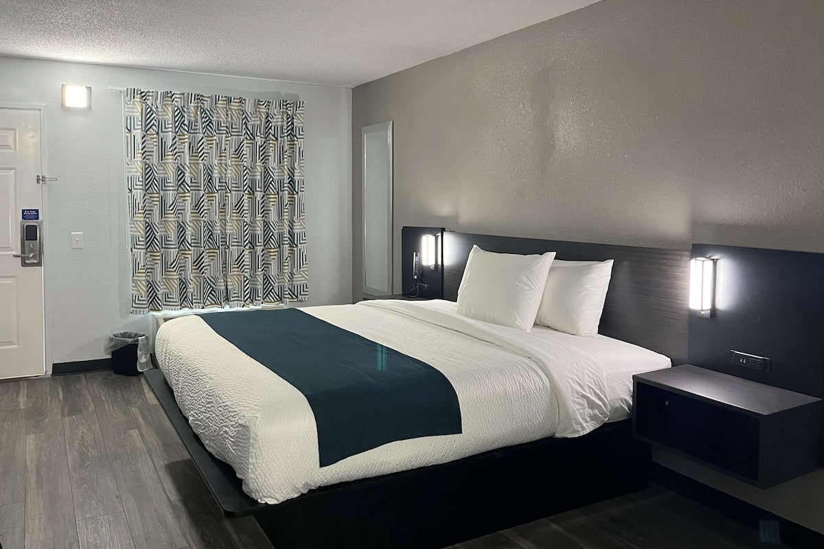 Une chambre d’hôtel moderne avec un grand lit, des tables de chevet, une tête de lit dotée d’un éclairage intégré, un rideau à motifs et un plancher en bois.