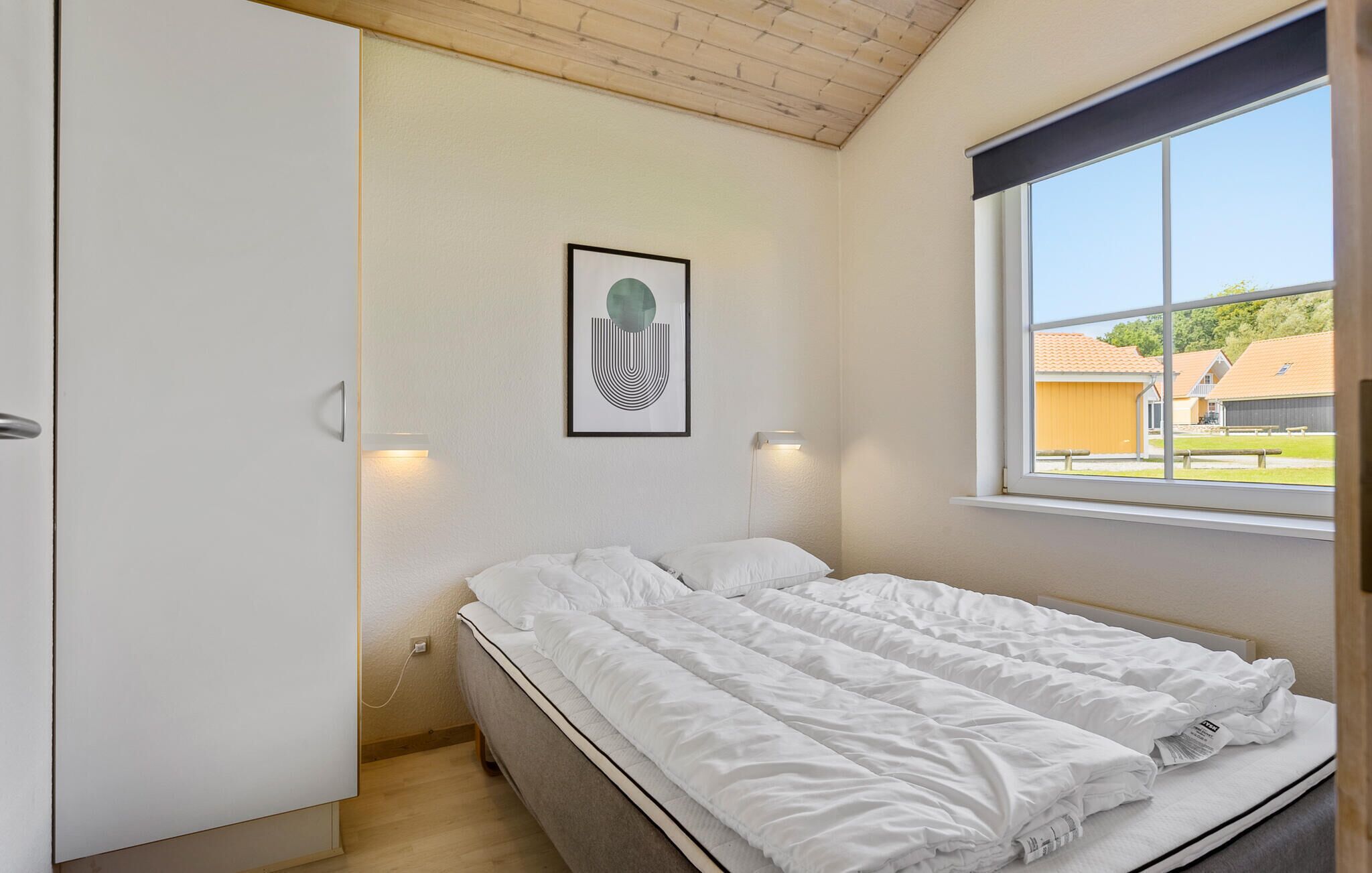 A bedroom with a bed, a wardrobe, a framed artwork, and a window with a view of buildings.