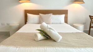 Una cama bien tendida con una toalla enrollada y una hoja verde, dos lámparas colgantes y un cabecero de madera en una habitación de hotel.