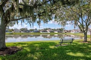A serene lakeside view with a bench, reflecting houses, and a tree with hanging moss. - Deer Run in Wesley Chapel 4br-3ba (Zephyrhills)