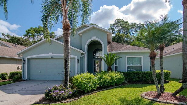A single-story house with a garage, surrounded by well-maintained landscaping including palm trees and flowering plants. - Deer Run in Wesley Chapel 4br-3ba (Zephyrhills)