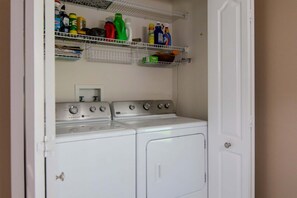 A laundry room with a washer and dryer, shelves above with cleaning supplies, and a white door. - Deer Run in Wesley Chapel 4br-3ba (Zephyrhills)