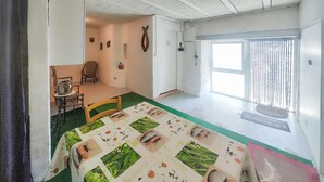 Una habitación con una cama cubierta por un edredón estampado con motivos florales, una mesita con una tetera, una silla y una ventana con cortinas.