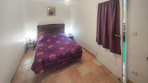 Un dormitorio con cama, mesita de noche, lámpara de pared, puerta con cortina y un ventanal pequeño.