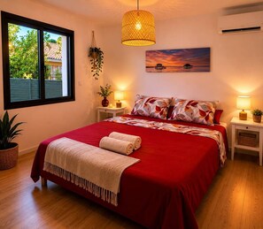 A bedroom with a large bed, two bedside tables, a hanging plant, a painting, and a window with a view.