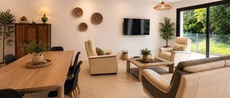 A modern living room with a wooden dining table, leather sofas, a flat-screen TV, and a large window with a view of greenery.