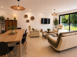 A modern living room with a wooden dining table, leather sofas, a flat-screen TV, and a large window with a view of greenery.