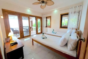 Ein Zimmer mit einem Bett, einem Schreibtisch mit Lampe und einem Deckenventilator. Es gibt Fenster und einen Blick ins Freie.