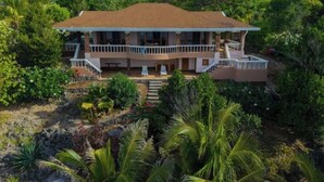 Une maison à deux étages dotée d’un balcon, entourée d’une végétation luxuriante et d’un paysage rocheux.