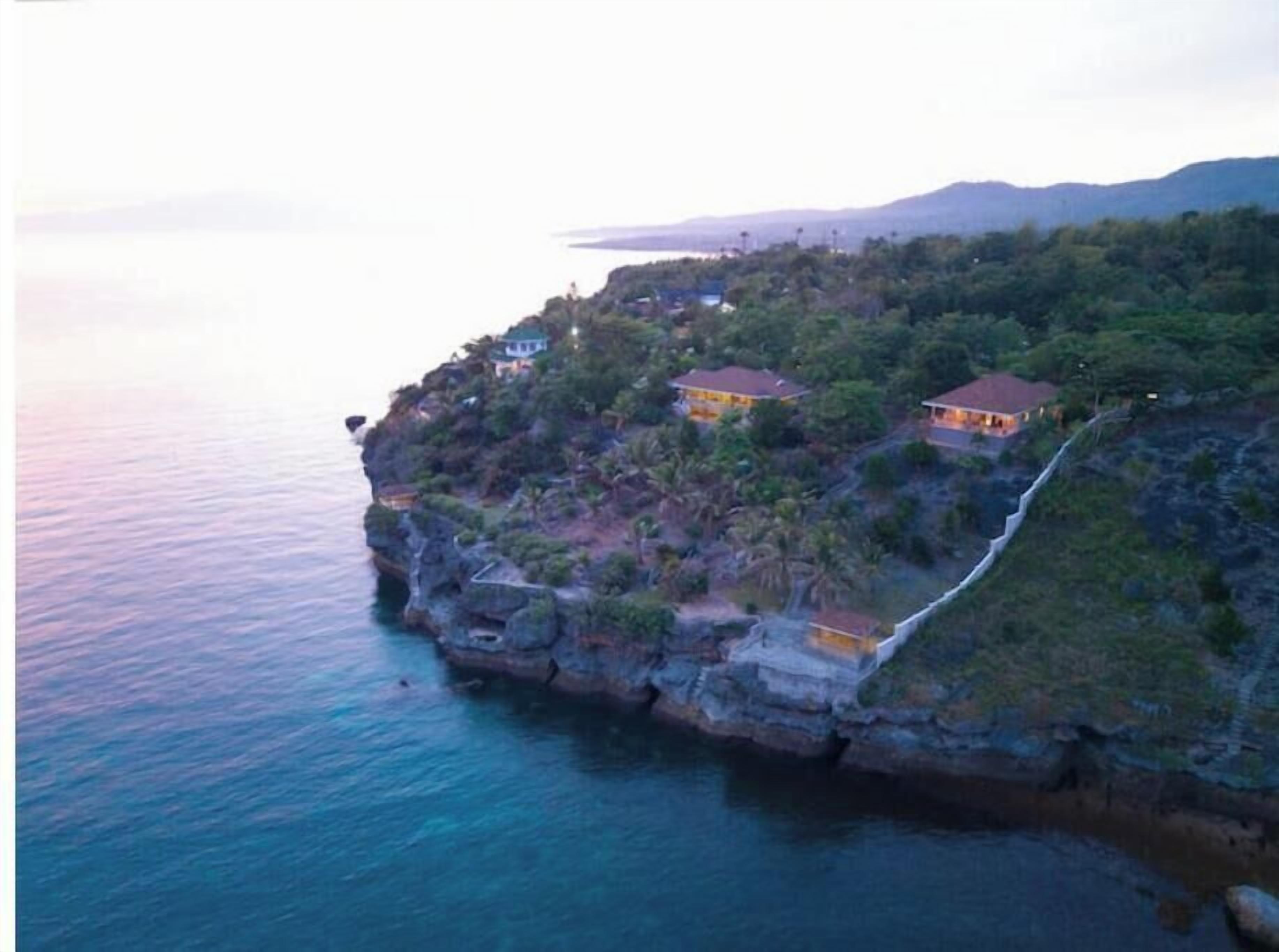 Une propriété située sur la côte, perchée sur une falaise, comprenant deux bâtiments aux toits rouges et un sentier menant à la mer.