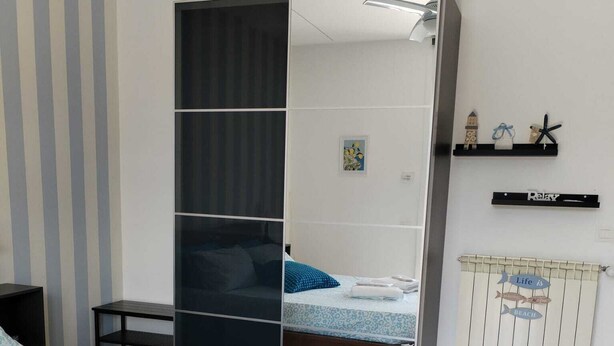 A bedroom with a bed, a wardrobe, a small table, and a shelf with decorative items. - Family apartment near Salerno, the Amalfi and Cilento coasts. (Baronissi)