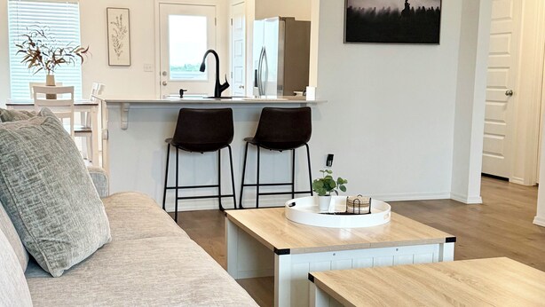 Una sala de estar moderna con un sofá, una mesa de centro y dos sillones. Se observa una zona de cocina con un refrigerador y un lavamanos. Un cuadro adorna la pared y se ve una puerta al fondo.