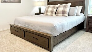 Un dormitorio con una cama de madera, ropa de cama blanca, una mesita de noche con una lámpara y un cuadro en la pared.