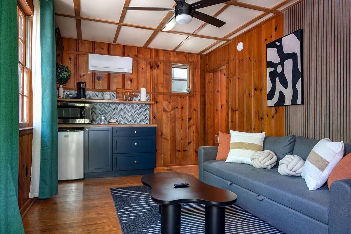 A cozy room with a kitchenette, a gray sofa with decorative pillows, a ceiling fan, and wooden walls.