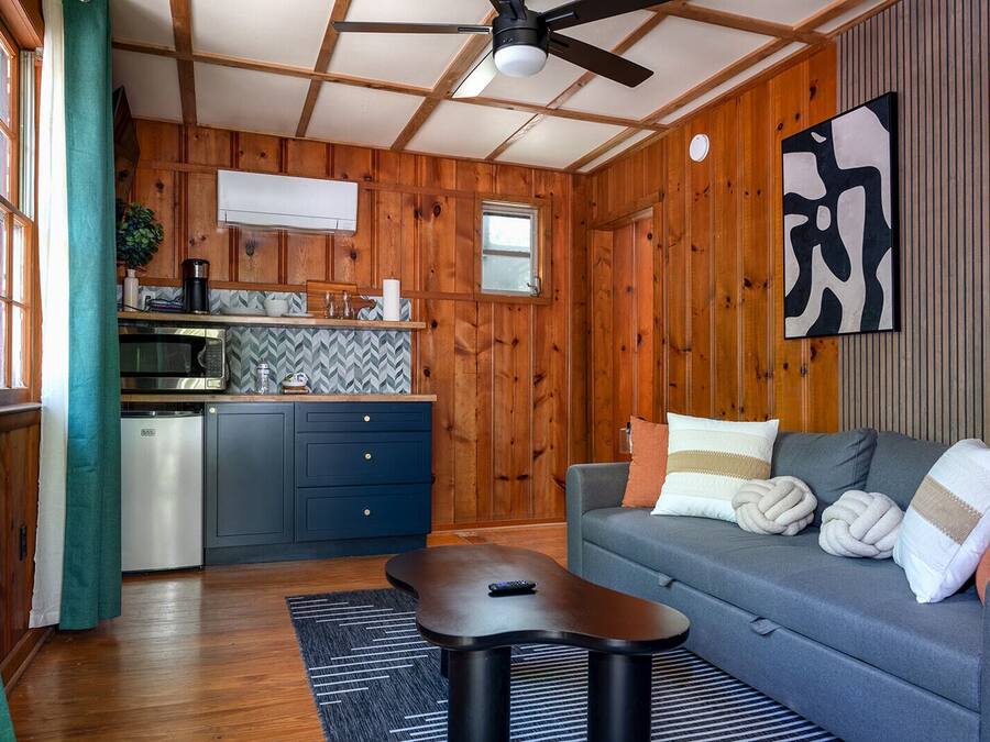 Um quarto acolhedor com cozinha compacta, um sofá cinza com almofadas decorativas, um ventilador de teto e paredes de madeira.