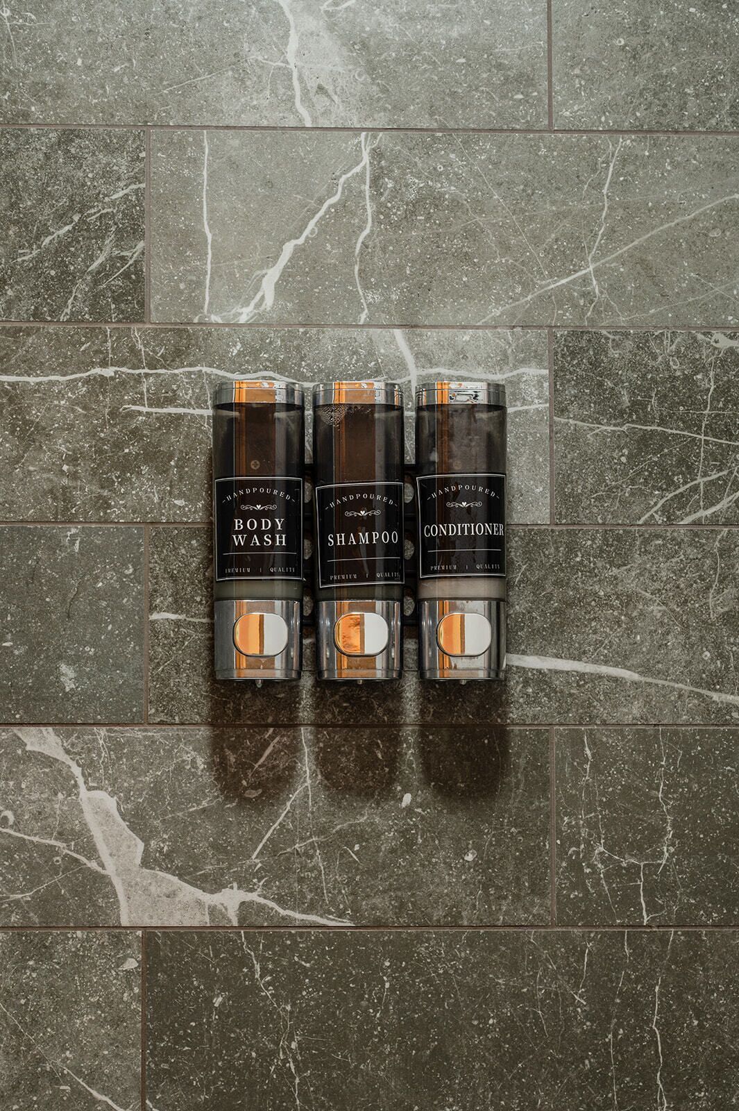 Three bottles of hand-poured body wash, shampoo, and conditioner on a marble floor.