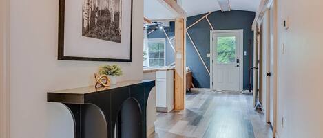 Un couloir avec un ventilateur de plafond, un tableau encadré, une plante posée sur une console et une porte donnant sur une autre pièce.