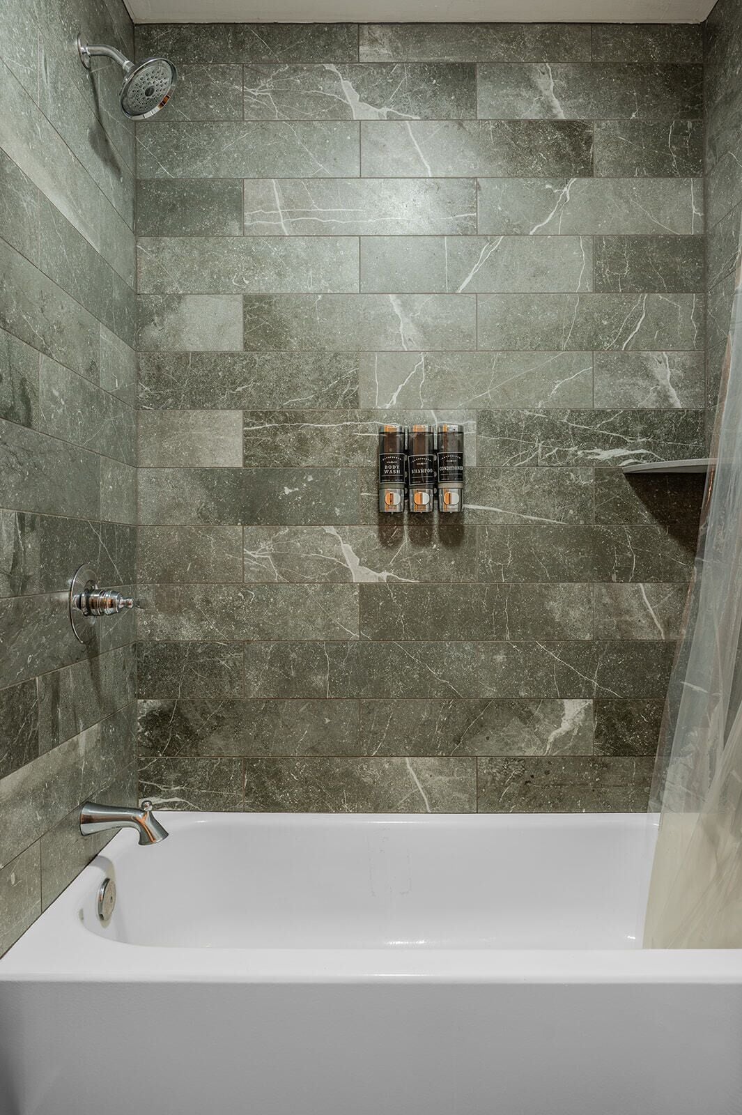A bathroom with a white bathtub, a showerhead, and three body wash bottles on the wall.