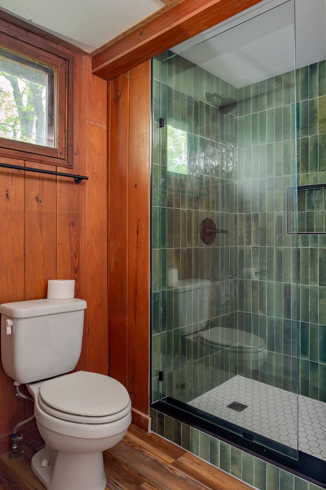 A bathroom with a toilet, wooden walls, a glass shower enclosure, and a window.