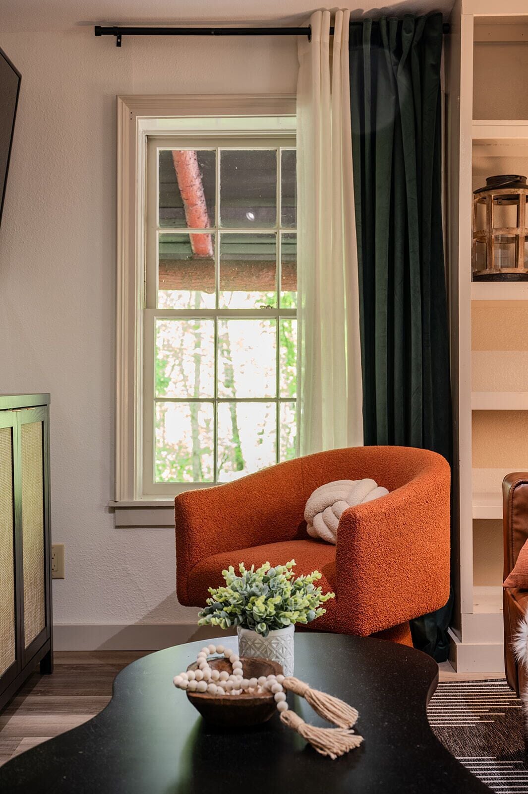A room with a window, a chair, a table with a plant and a beaded necklace, and a bookshelf.