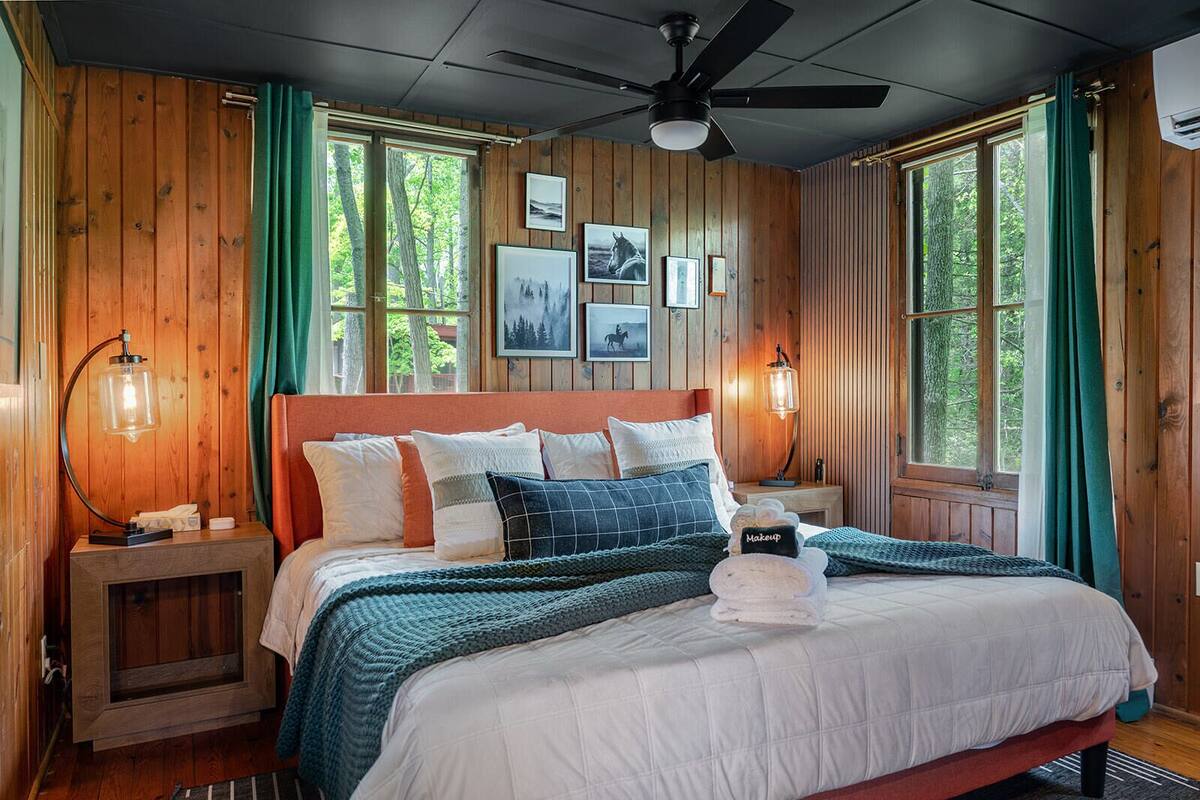A wooden-paneled bedroom with a bed, framed pictures, a ceiling fan, and a nightstand with a lamp.