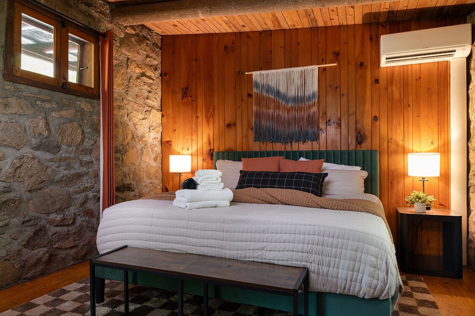 A bedroom with a stone wall, wooden paneling, a bed with white bedding, a bench, two bedside tables with lamps, and a hanging tapestry.