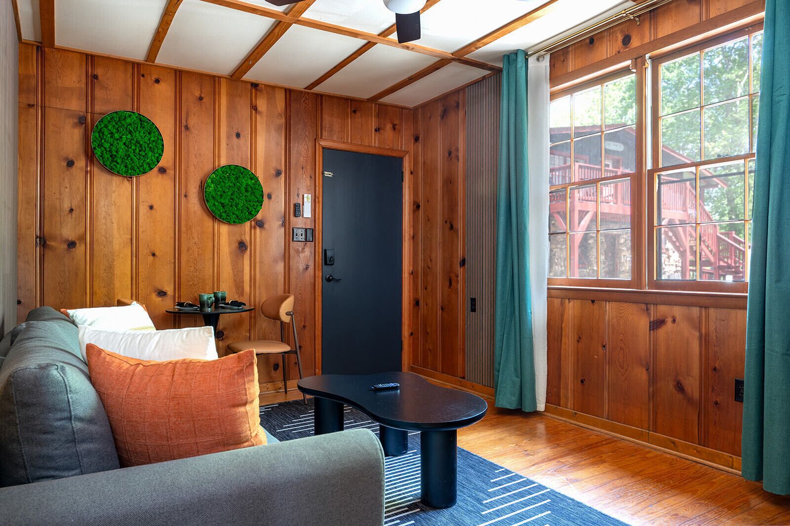 A room with wooden walls, a black door, a sofa, a table, and a window with curtains.