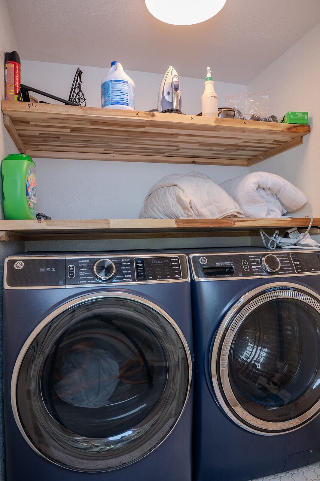 A laundry area with two front-loading washing machines, a shelf with cleaning supplies, and a bed with pillows.