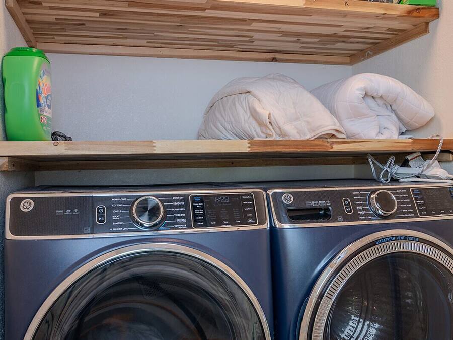 A laundry area with two front-loading washing machines, a shelf with cleaning supplies, and a bed with pillows.