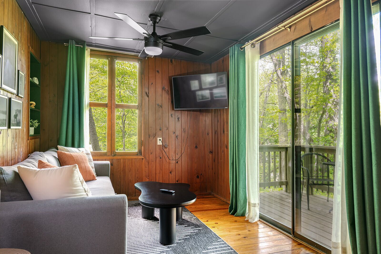 Une pièce dotée d’un ventilateur de plafond, d’un téléviseur fixé au mur, d’un canapé avec des coussins, d’une petite table et d’une porte-fenêtre coulissante donnant sur une terrasse extérieure.