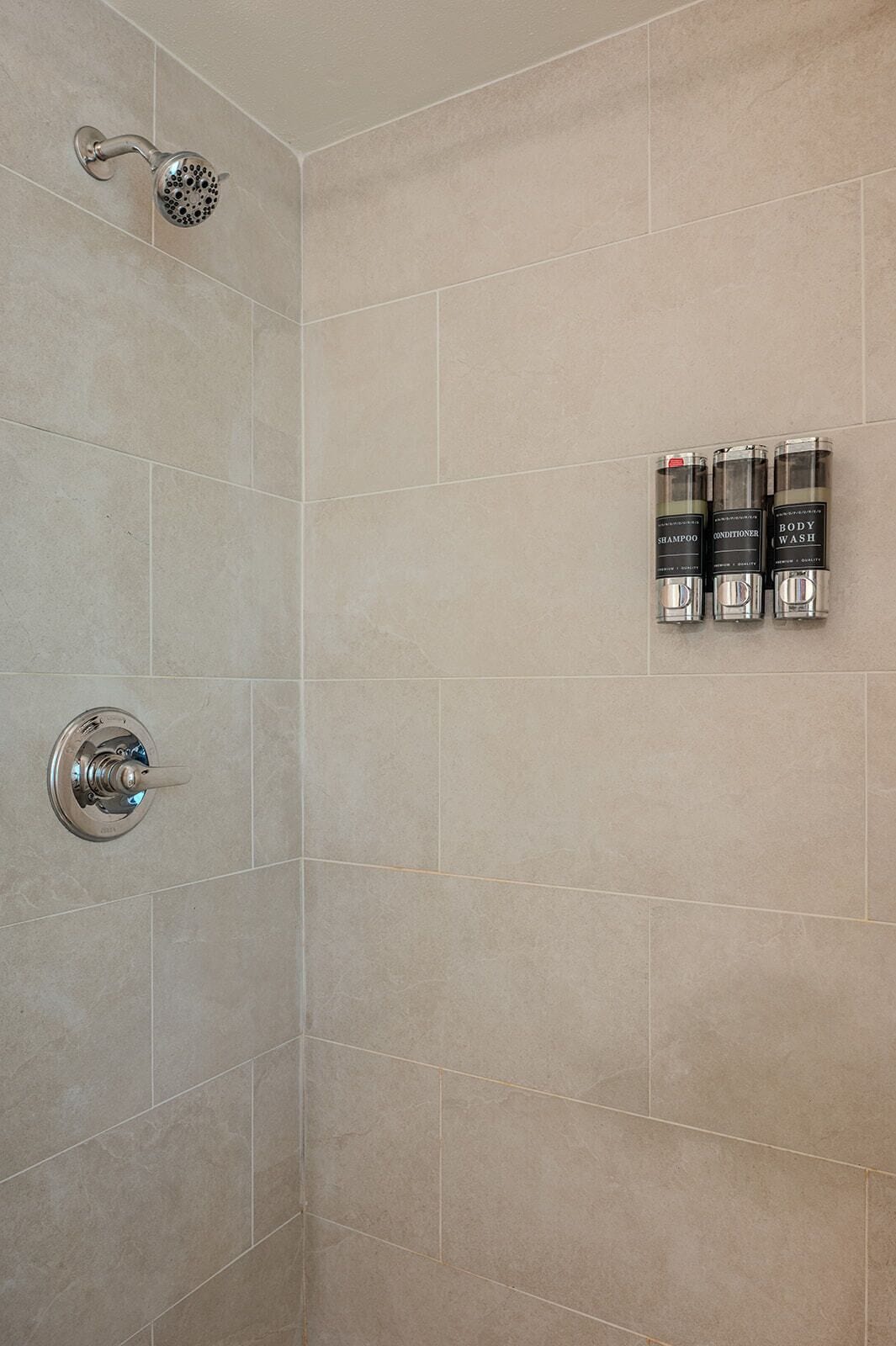 A shower with a handheld showerhead and a wall-mounted control valve. There are three bottles of body wash, shampoo, and conditioner on the wall.