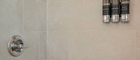 A shower with a handheld showerhead and a wall-mounted control valve. There are three bottles of body wash, shampoo, and conditioner on the wall.