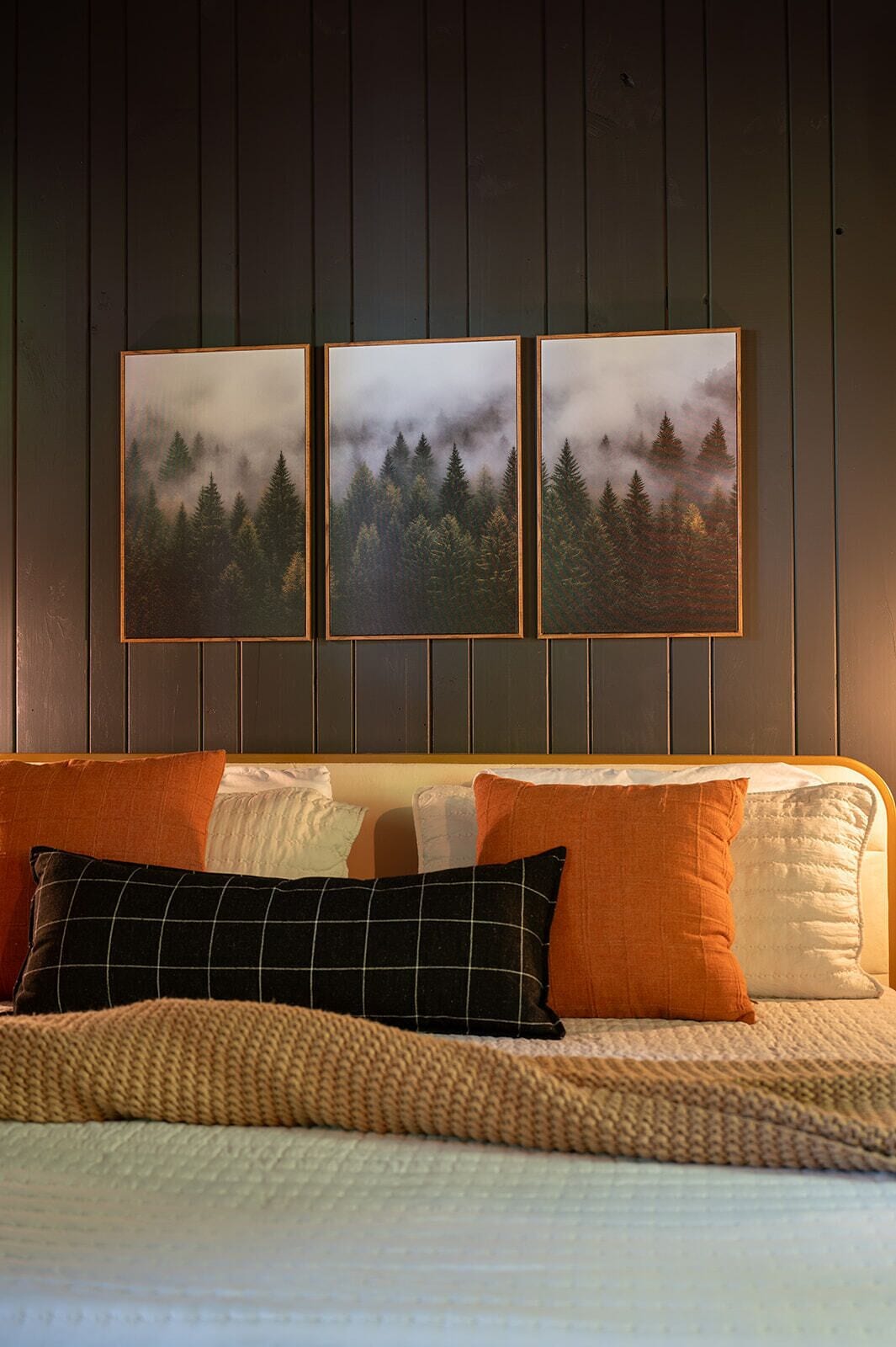 Ein Bett mit einer gewebten Decke, einem Kopfteil und drei gerahmten Bildern eines Waldes an der Wand.