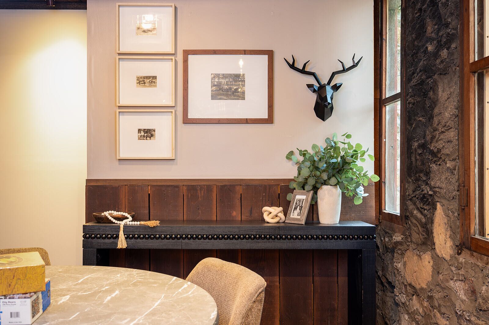 A room with a wooden bench, a marble table, framed pictures, a deer head decoration, and a plant.